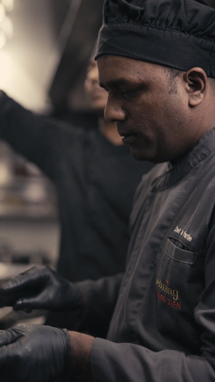 Chef wearing black uniform and gloves preparing food in a busy kitchen.